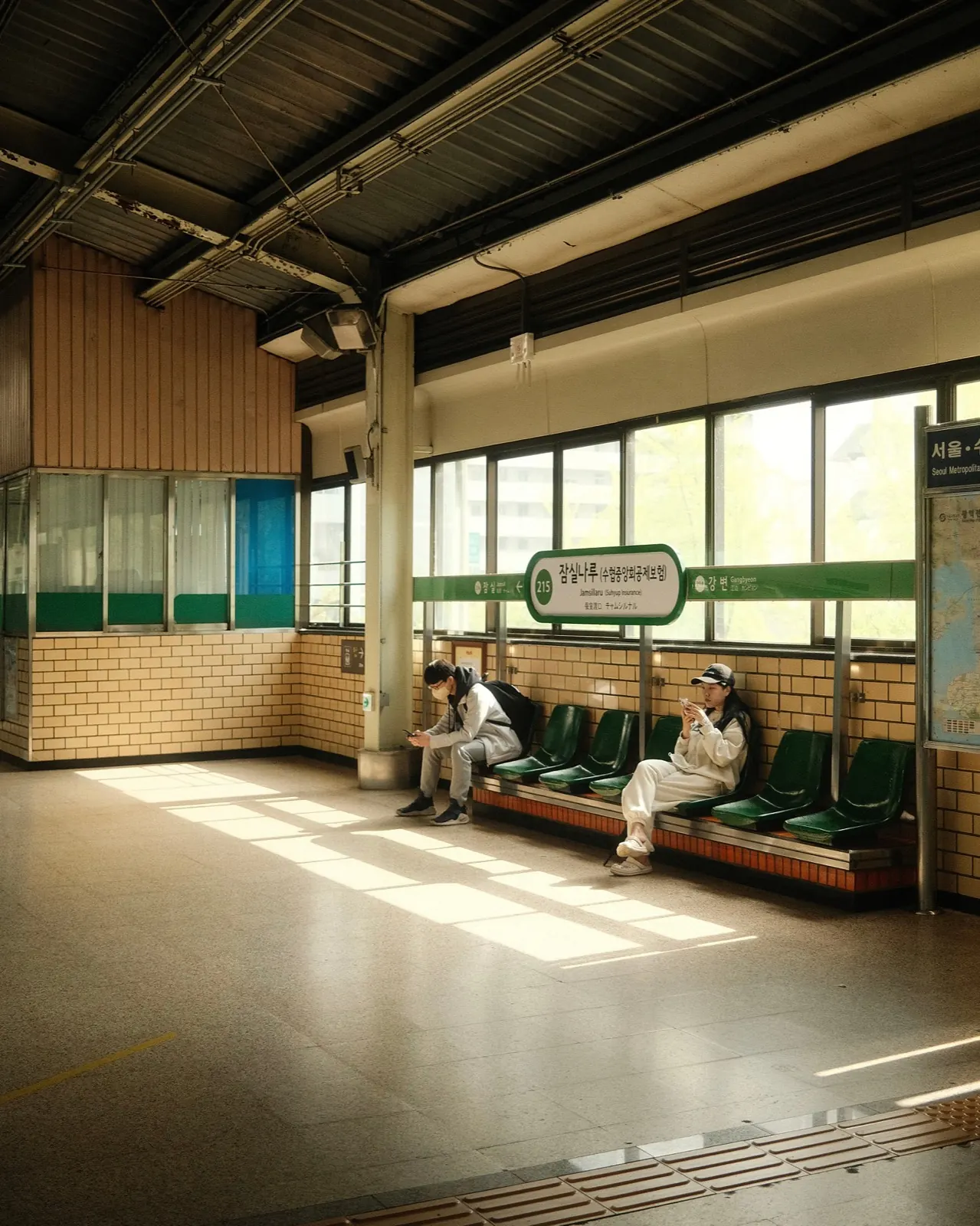 Two commuters waiting on a bench in the afternoon light at Jamsillaru Station, Seoul