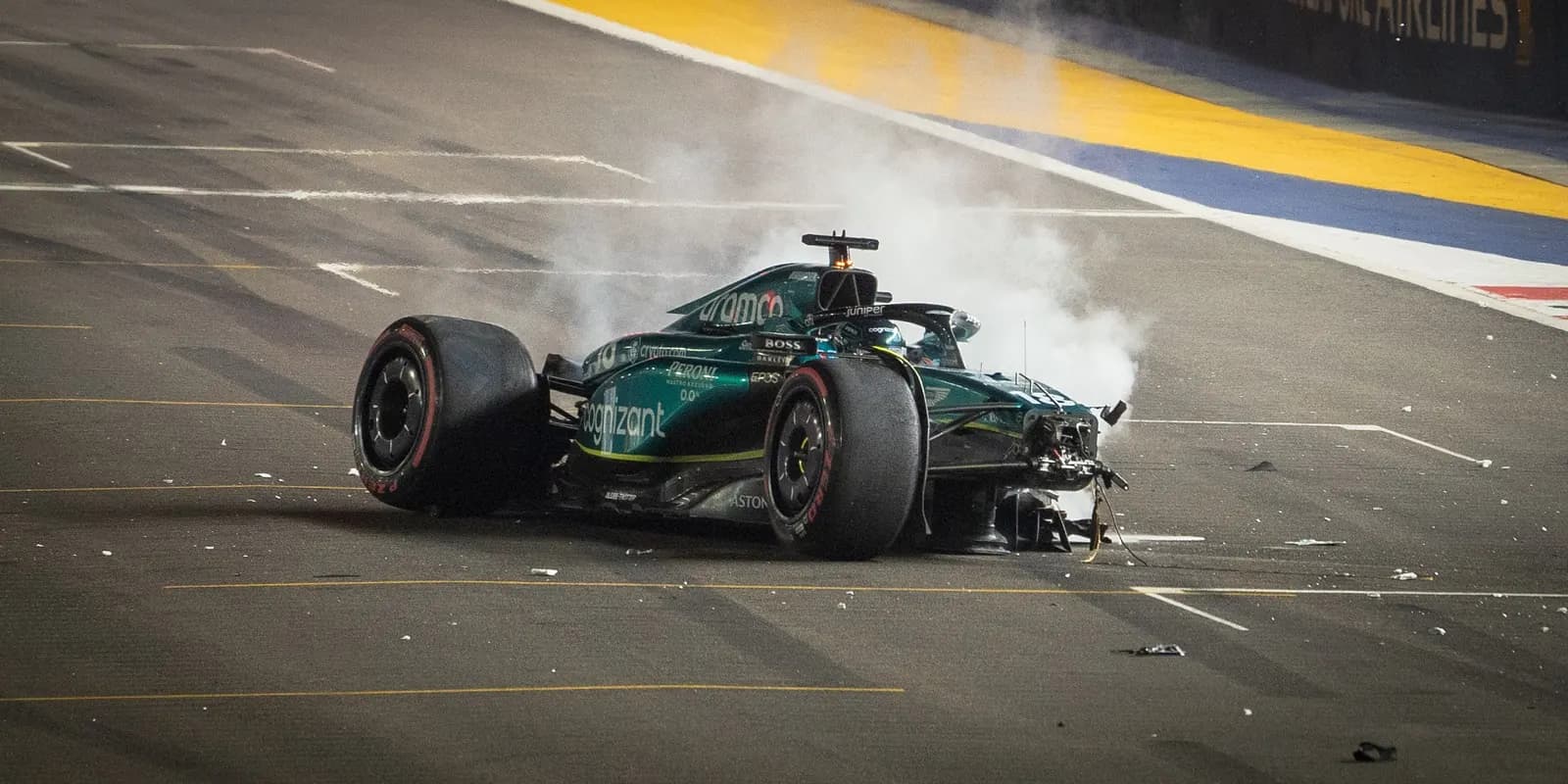 An Aston Martin wrecked on track under floodlights, smoking, front wing torn away and debris scattered across the asphalt